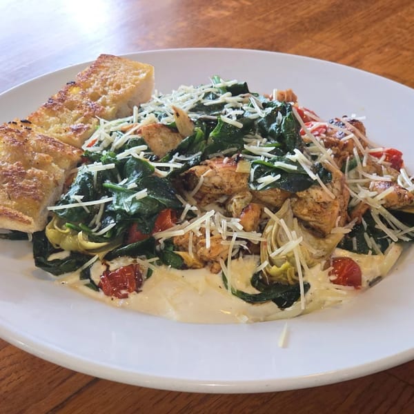 Creamy chicken pasta with spinach, tomatoes, artichokes, and garlic bread on a white plate.
