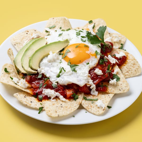 Nachos with fried egg, avocado slices, red salsa, and crumbled cheese on a white plate.